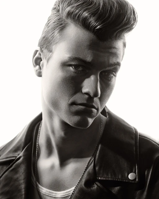 Monochrome portrait of a handsome adult male with pompadour hairstyle, wearing a leather jacket and chain necklace, against a white backdrop.